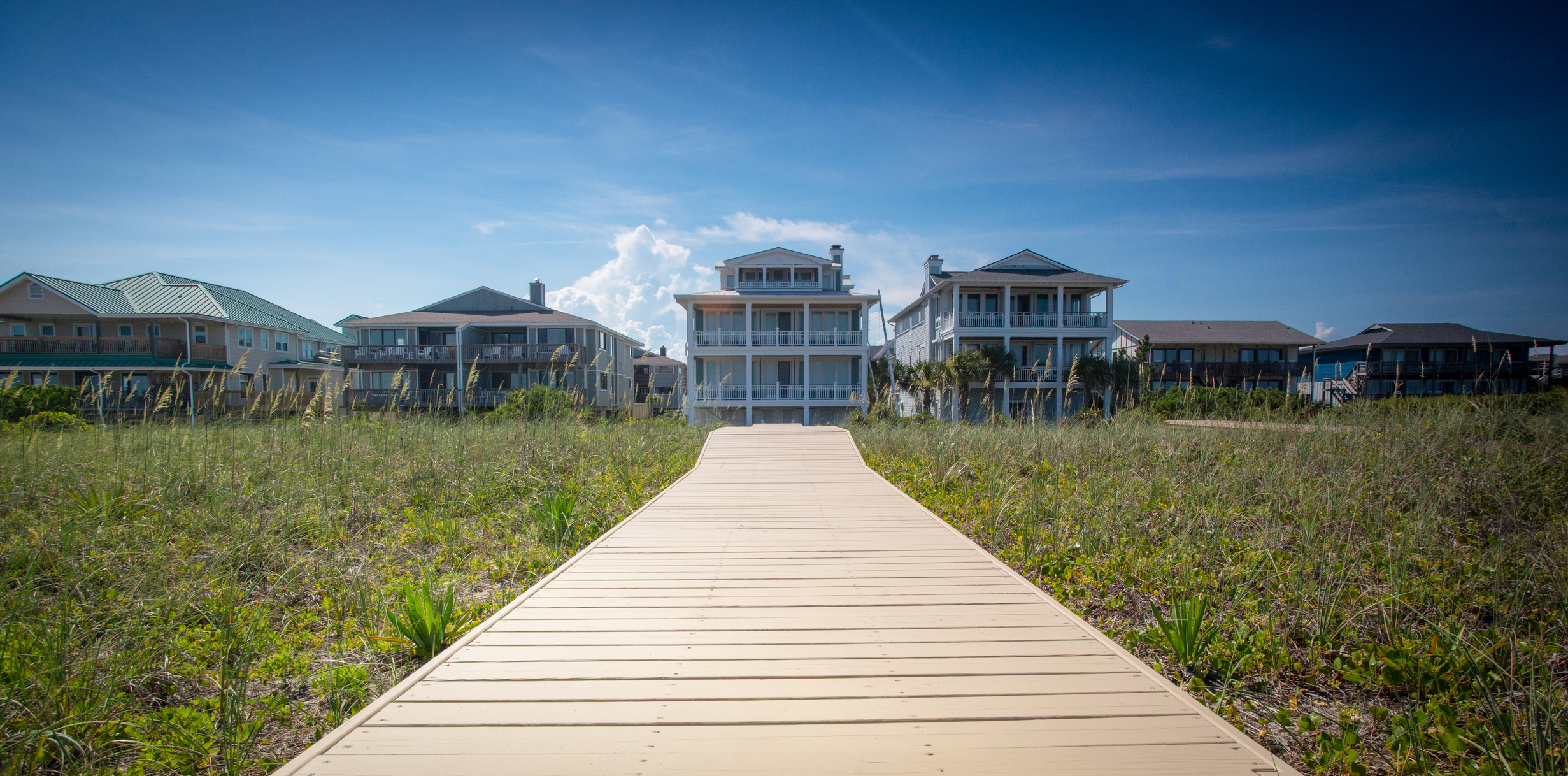 A long wood plank walkway leads to a large beach house.